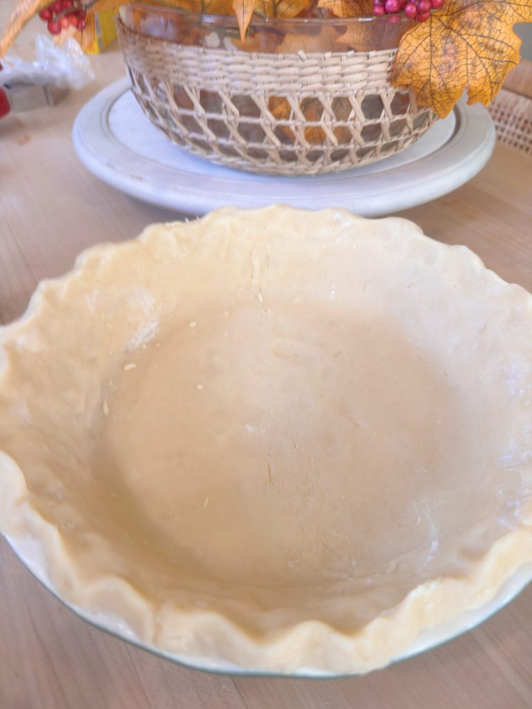 An unbaked pie crust in a glass pie dish sits on a wooden surface, with a fall-themed centerpiece in the background.
