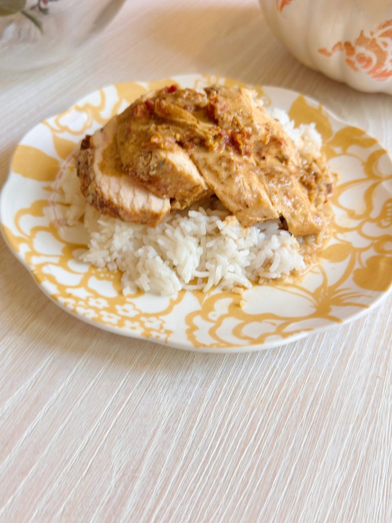 A plate with white rice topped with sliced chicken breast and creamy sauce, served on a patterned dish, sits on a light-colored table.