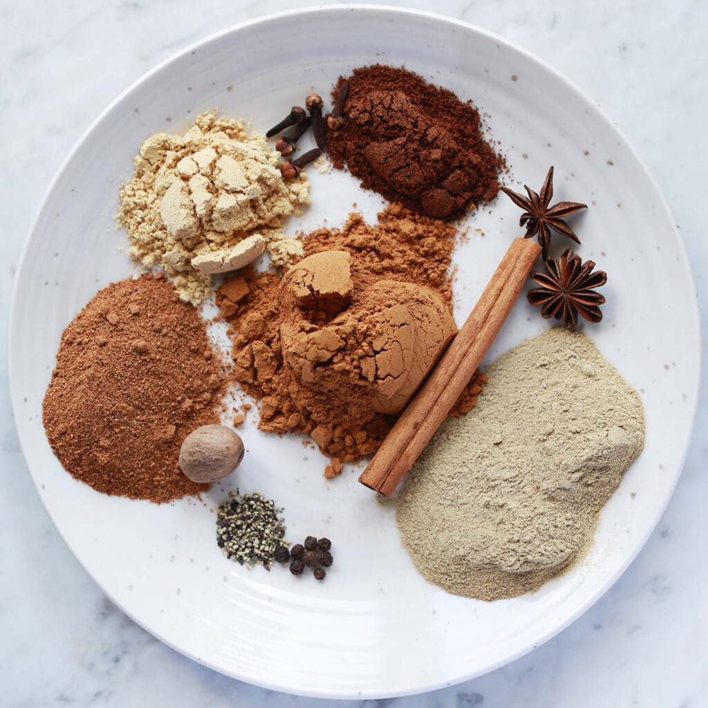 A white plate with piles of various ground spices, including cinnamon, ginger, nutmeg, cloves, star anise, and black pepper, arranged separately on a marble surface.