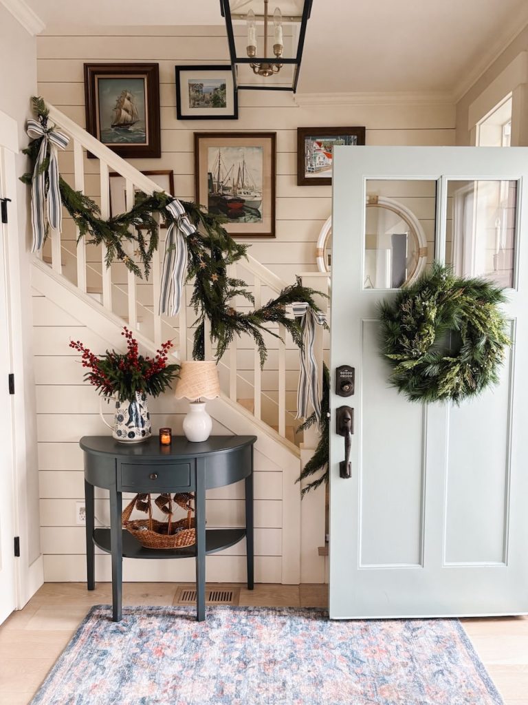 Entryway with a light blue door decorated with a wreath, staircase with garland and ribbons, framed art on the wall, a table with a lamp, vase, basket, and a patterned rug.