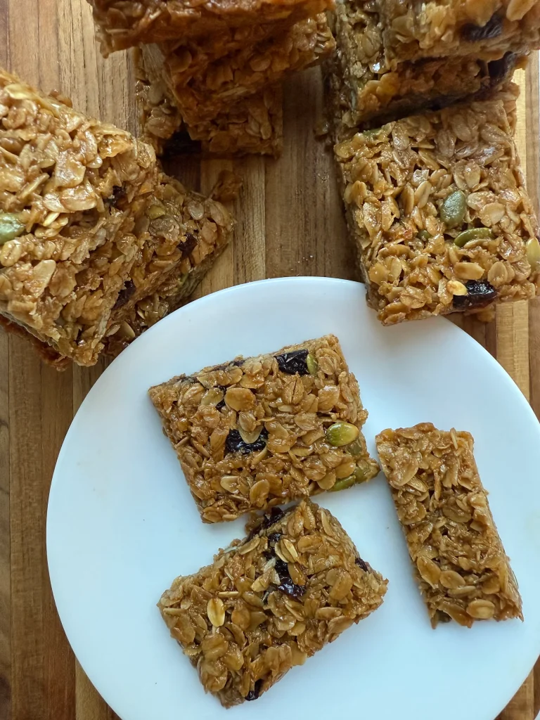 Granola bars with oats, seeds, and dried fruit are arranged on a wooden surface, with three bars placed on a white plate.