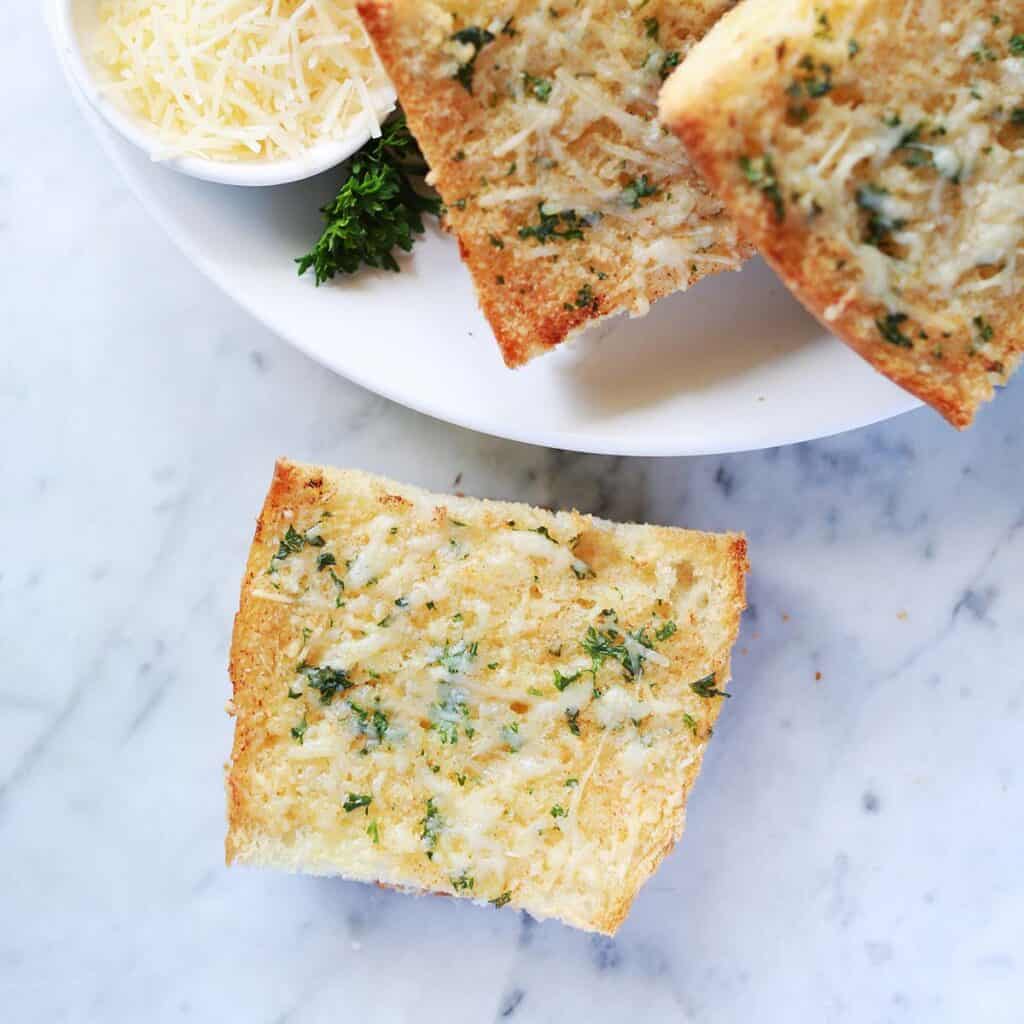 Slices of garlic bread topped with melted cheese and herbs on a marble surface, with a bowl of shredded cheese on the side.