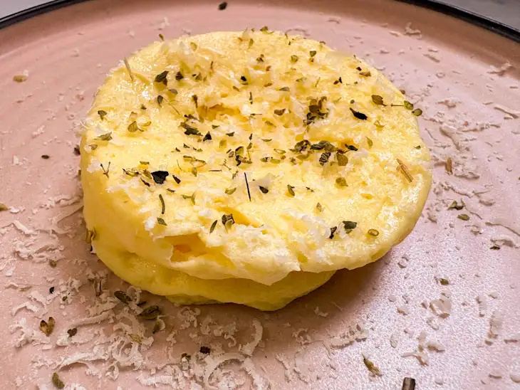A fluffy yellow omelette garnished with grated cheese and dried herbs sits on a beige plate.