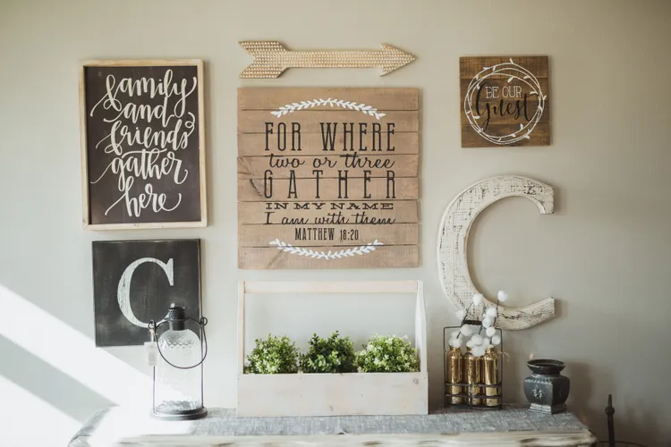 A farmhouse-style wall display featuring framed quotes, a large wooden "C," a decorative arrow, faux plants, cotton stems, and a lantern on a light-colored console table.
