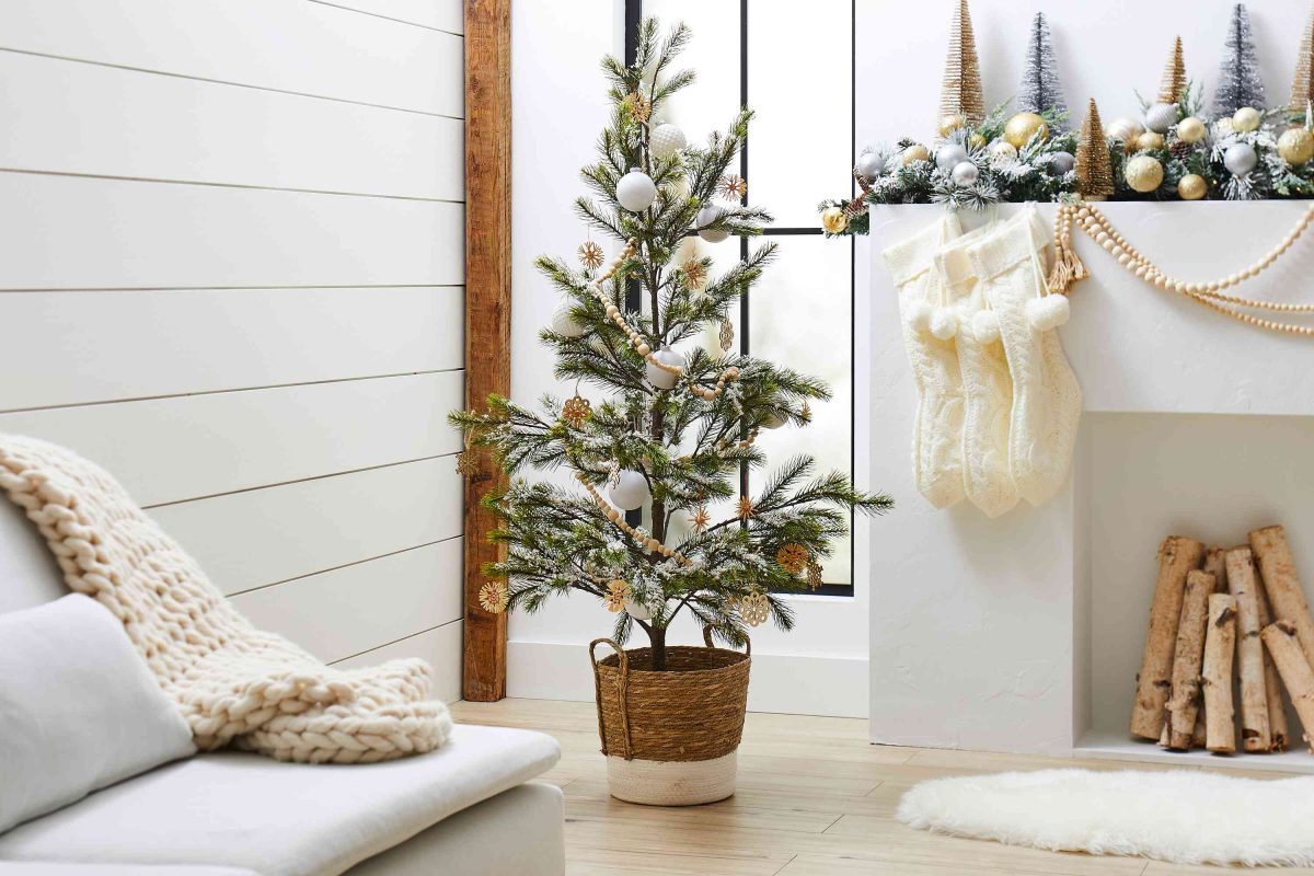 A small decorated Christmas tree in a wicker basket stands next to a fireplace with stockings, festive garland, and neatly stacked firewood in a bright living room.