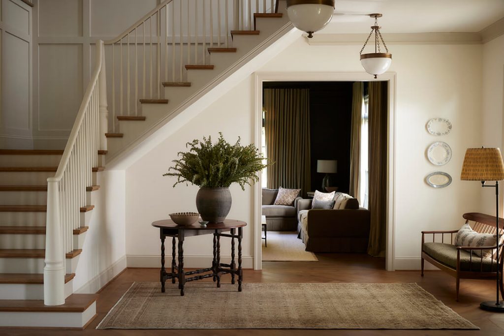 A well-lit entryway with a staircase, a round table with a large vase of greenery, a bench, and a view into a cozy living room with sofas and curtains.