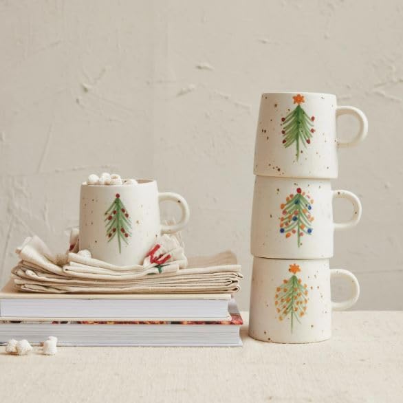 Four ceramic mugs with Christmas tree designs; three are stacked, and one sits on a folded napkin atop books, filled with a drink and marshmallows.