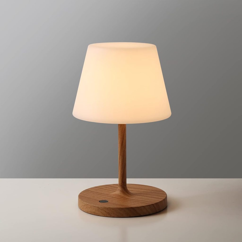 A small modern table lamp with a wooden base and stem, topped by a white, conical lampshade, sits on a plain white surface against a gray background.