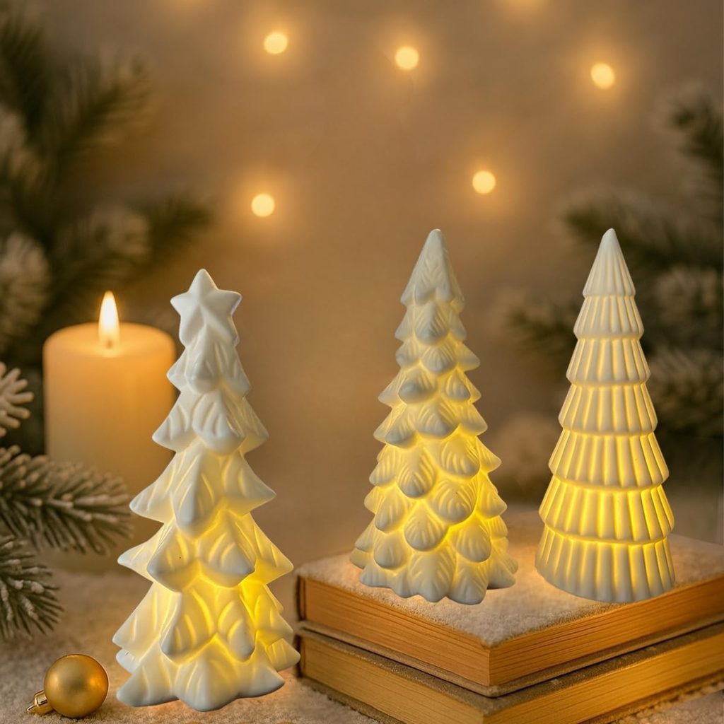 Three white ceramic Christmas tree decorations with warm lights inside are displayed on books, with a lit candle, pine branches, and string lights in the background.