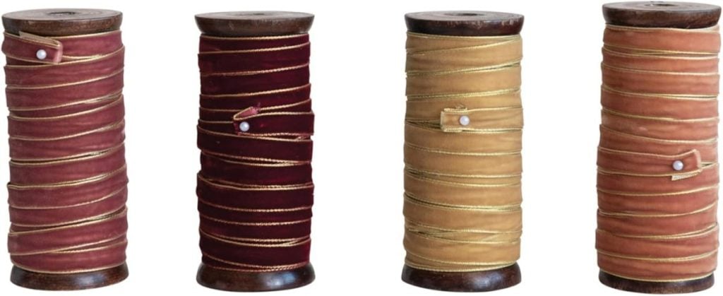 Four large wooden spools are wrapped with velvet ribbon in varying shades of red and gold, each secured with a pearl-headed pin.