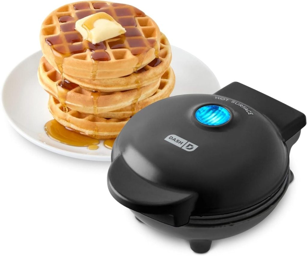 A stack of three waffles topped with butter and syrup on a white plate sits next to a closed black Dash mini waffle maker.