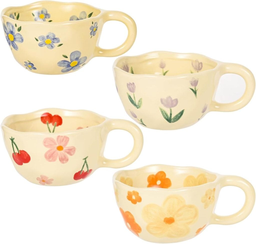 Four cream-colored ceramic mugs with scalloped rims, each featuring a different hand-painted floral or fruit design in various colors.