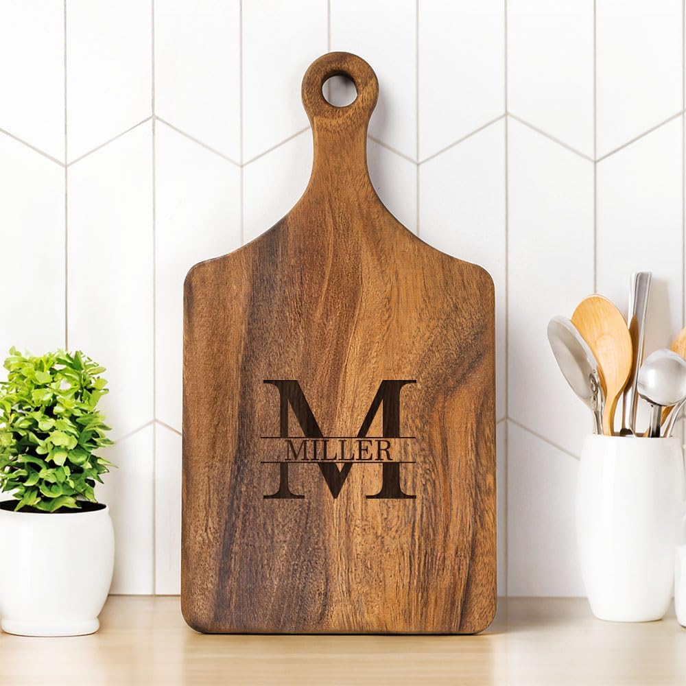 A wooden cutting board with a handle and the name "Miller" engraved over a large letter "M," standing upright on a kitchen counter beside utensils and a small potted plant.