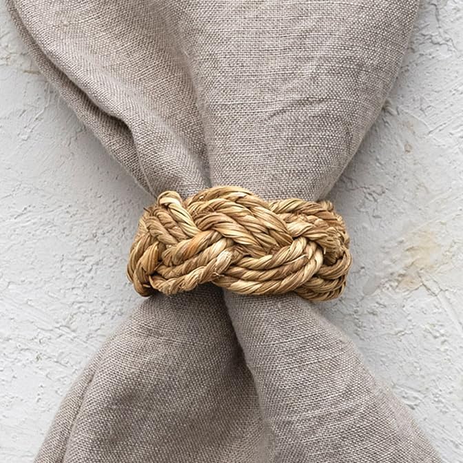 A beige linen napkin is held by a woven straw napkin ring against a textured white background.