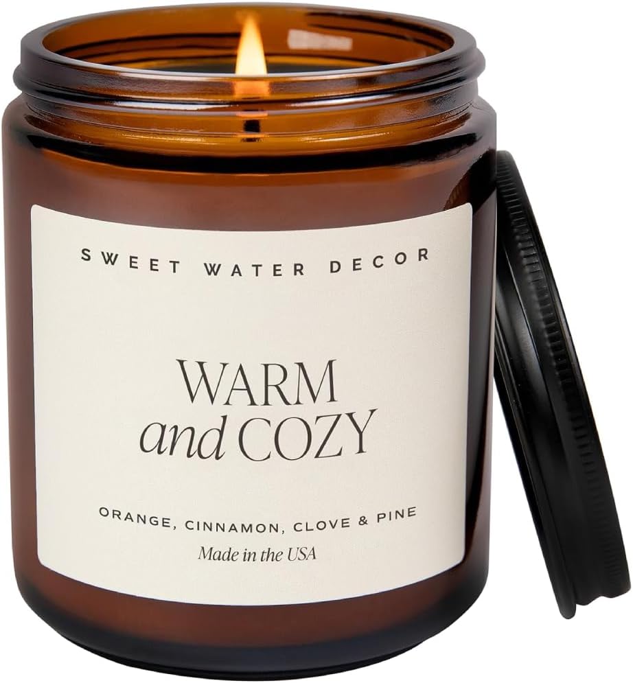 A lit brown glass jar candle labeled "Warm and Cozy" with notes of orange, cinnamon, clove, and pine; the lid rests beside the jar.