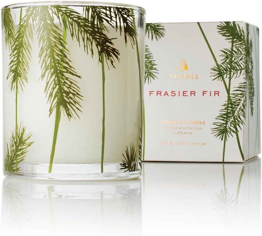 A glass candle decorated with green fir branches sits in front of a matching box labeled "Thymes Frasier Fir" with gold text and accents.