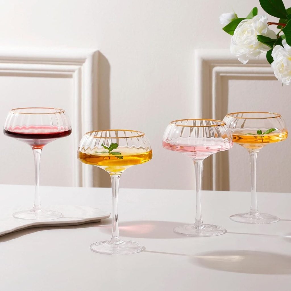 Four elegant coupe glasses with different colored drinks and garnishes sit on a white surface next to a white flower arrangement against a light background.