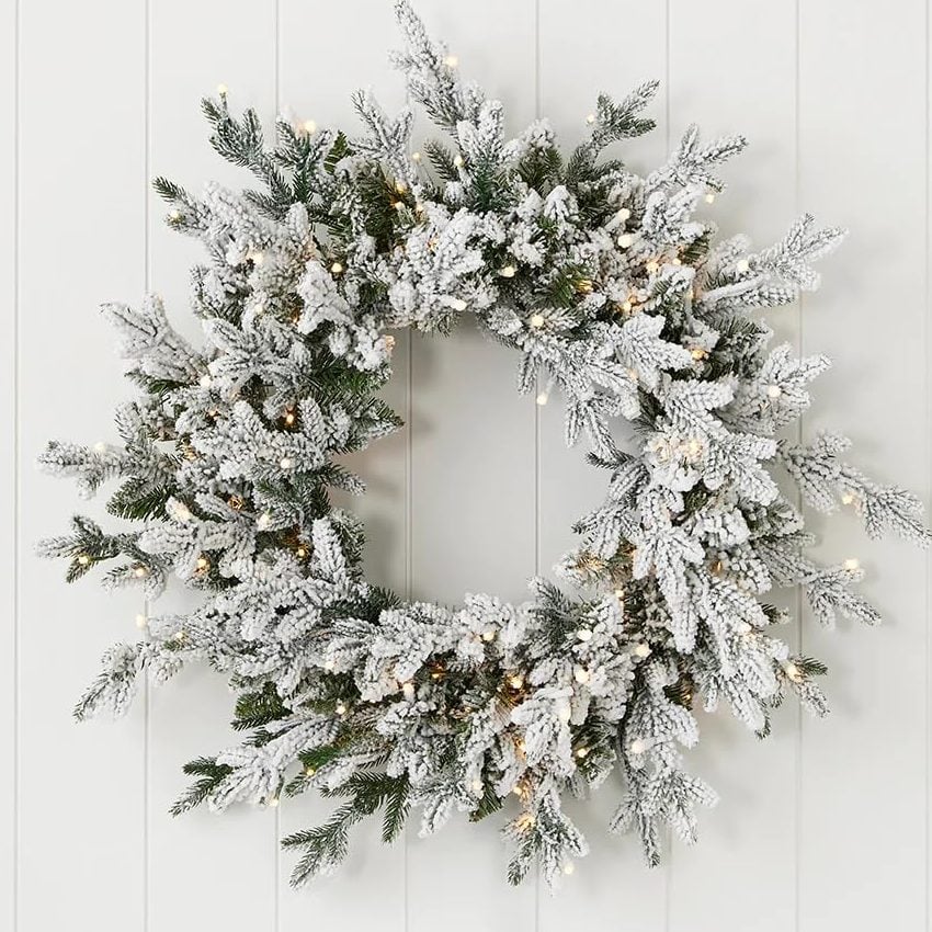 A round, snow-frosted artificial pine wreath with small white lights hangs on a white paneled wall, creating window art that brightens any space with festive charm.