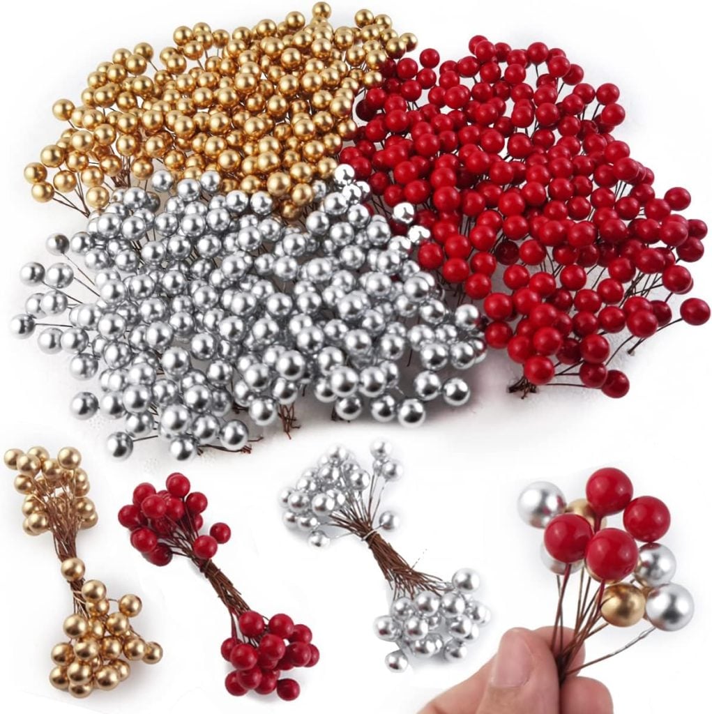Assorted clusters of artificial berries in metallic gold, red, and silver colors arranged on a white background. A hand holds a bunch of the berries in the foreground.