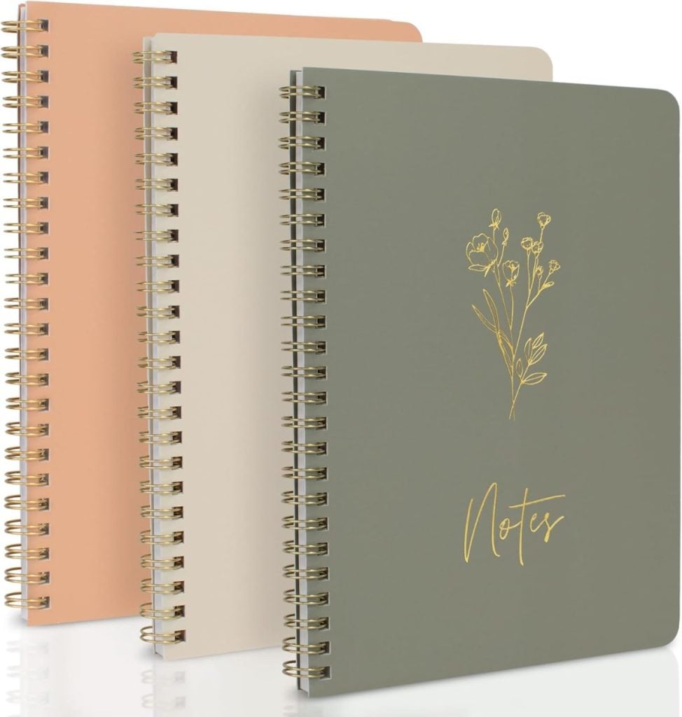 Three spiral-bound notebooks in pastel colors are stacked, with the front cover featuring a gold floral illustration and the word "Notes.