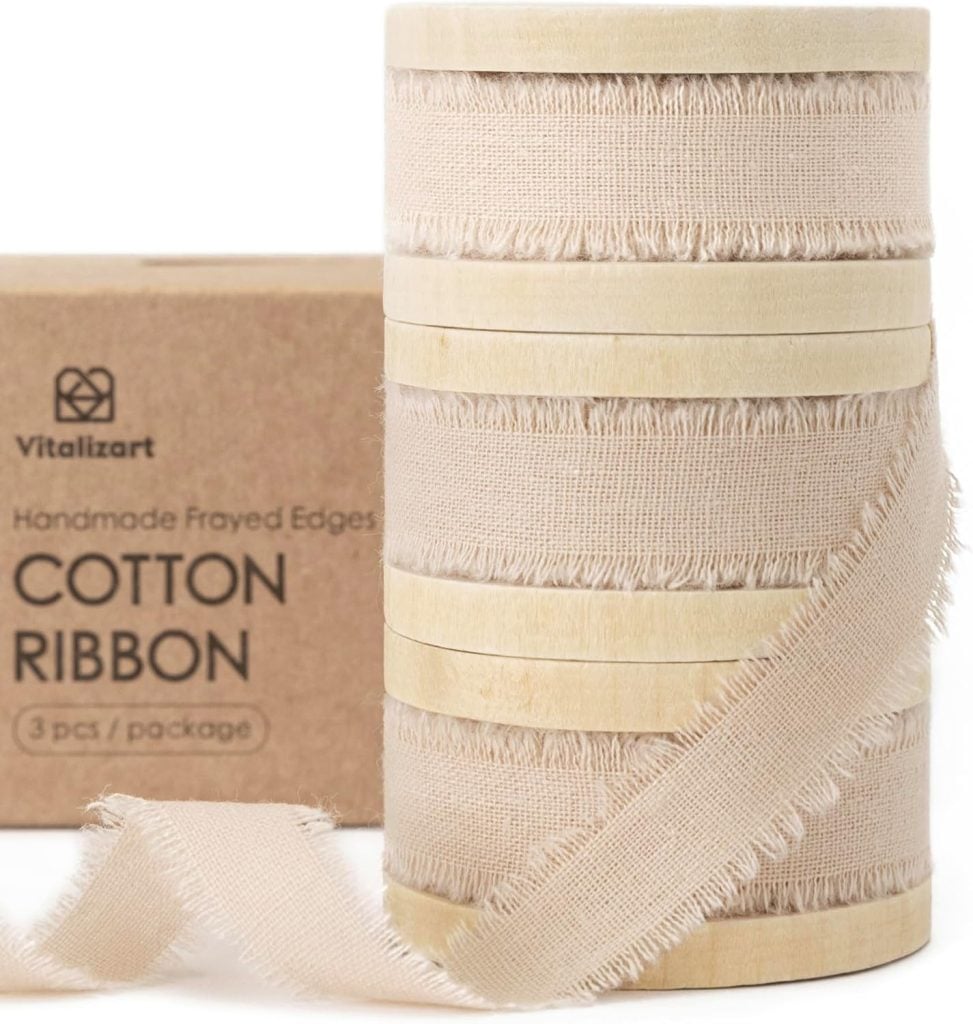 A spool of beige cotton ribbon with frayed edges is shown beside a cardboard box labeled "Vitalizart Handmade Frayed Edges Cotton Ribbon, 3 pcs/package.