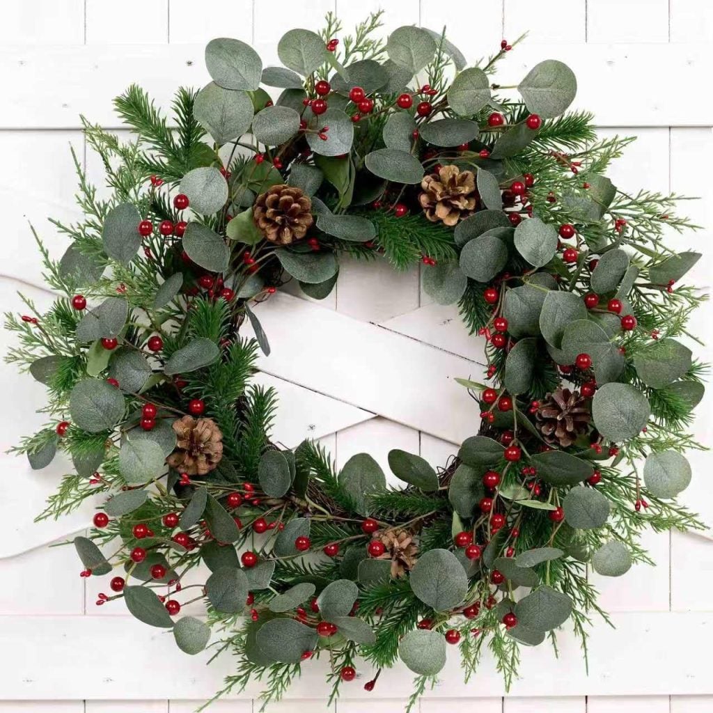 A decorative wreath made of green leaves, pinecones, and red berries is hung on a white wooden door, creating a festive touch reminiscent of window art.