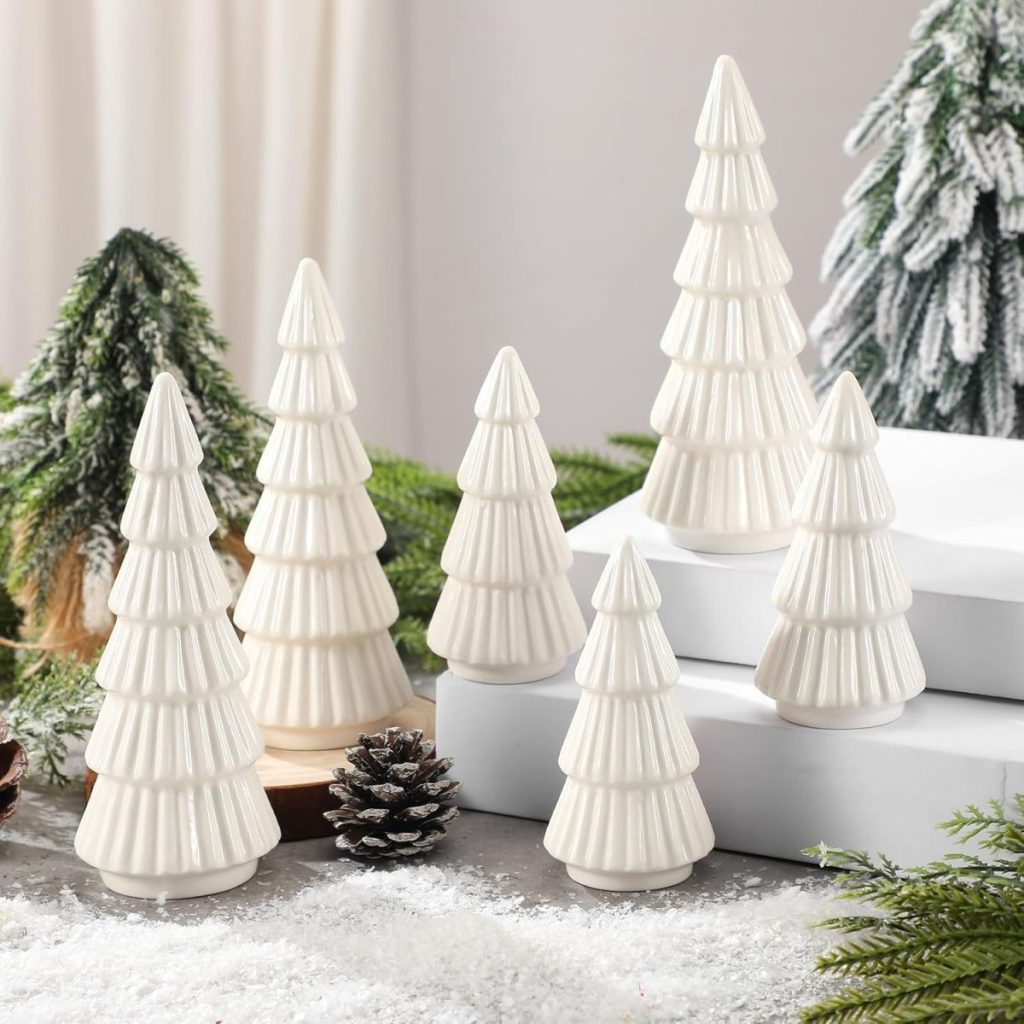Six white ceramic Christmas tree figurines are arranged on a snowy surface, surrounded by artificial greenery, a pine cone, and snow-dusted decorations.