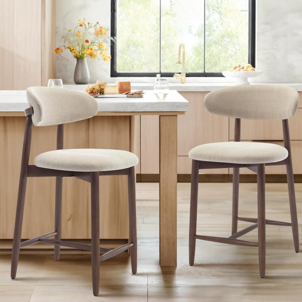 Two upholstered barstools with wooden legs are placed in front of a kitchen island in a modern, light-filled kitchen with neutral tones and minimal decor—perfect for enjoying drinks or showcasing a thoughtful hostess gift.