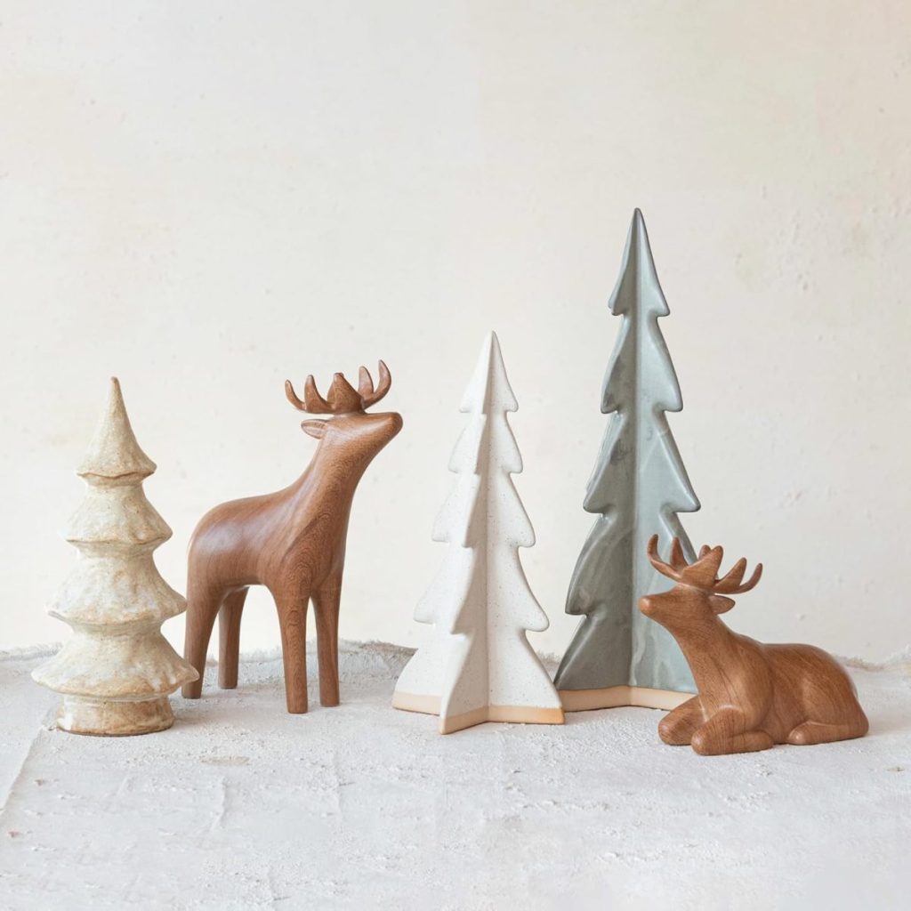 Wooden and ceramic figurines of deer and evergreen trees arranged on a neutral surface against a light background.