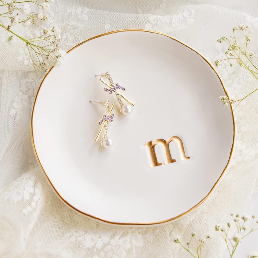 A white plate with gold trim holds a pair of gold and pearl earrings and a gold letter "m," set on a lace fabric with small white flowers.