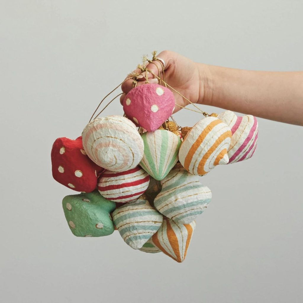 A hand holds a bunch of colorful, striped and polka-dotted paper ornaments shaped like seashells against a plain background.