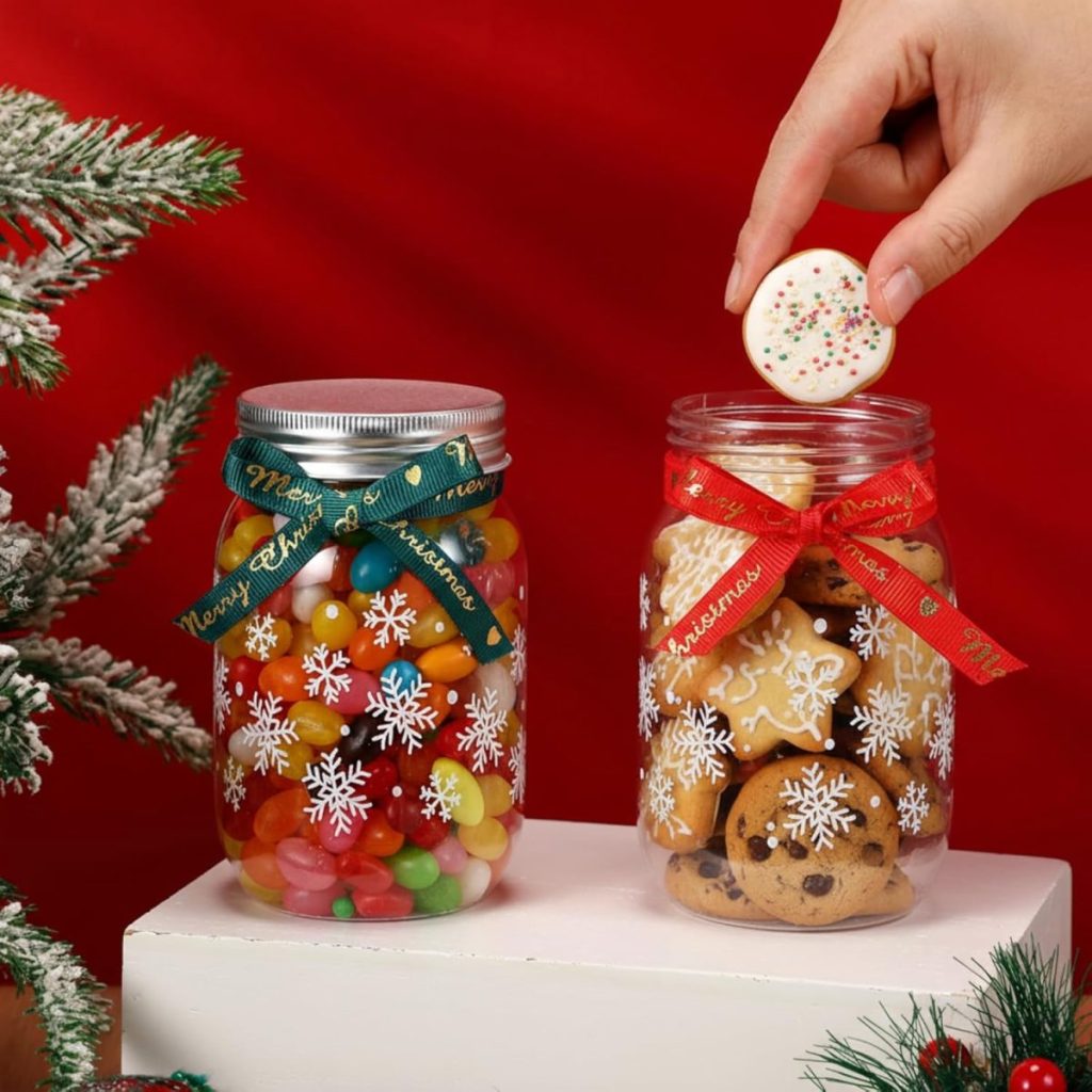 Two glass jars decorated with snowflakes and ribbons—one filled with colorful jellybeans, the other with cookies. A hand is placing a cookie into the jar beside a frosted pine branch—perfect as a festive hostess gift.