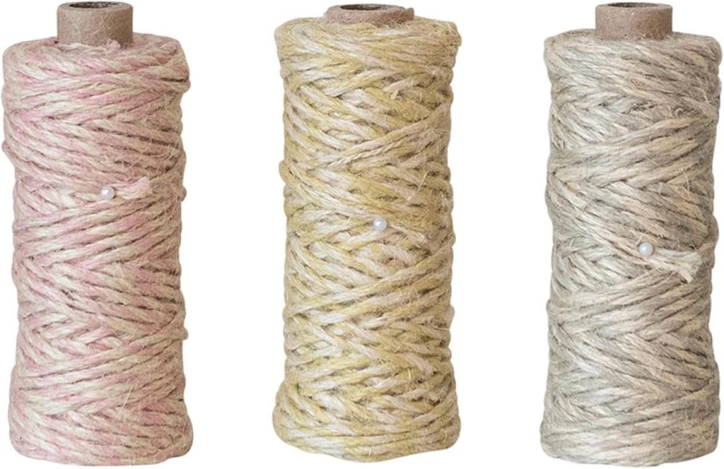Three spools of twine in pastel pink, yellow, and beige colors, each wrapped with a small pearl-headed pin.