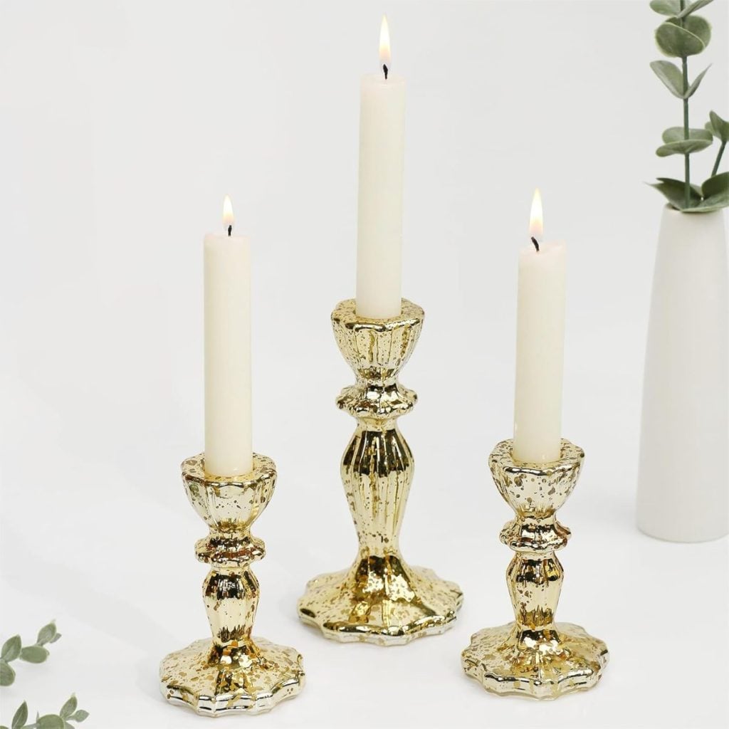 Three gold-colored candle holders of varying heights, each holding a lit white taper candle, are arranged on a white surface with small green plants nearby.