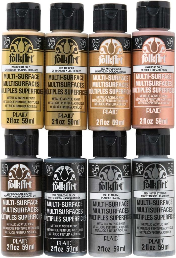 Nine bottles of FolkArt multi-surface acrylic paint in various metallic and neutral shades are arranged in a three-by-three grid. Each bottle contains 2 fl oz (59 ml) of paint.