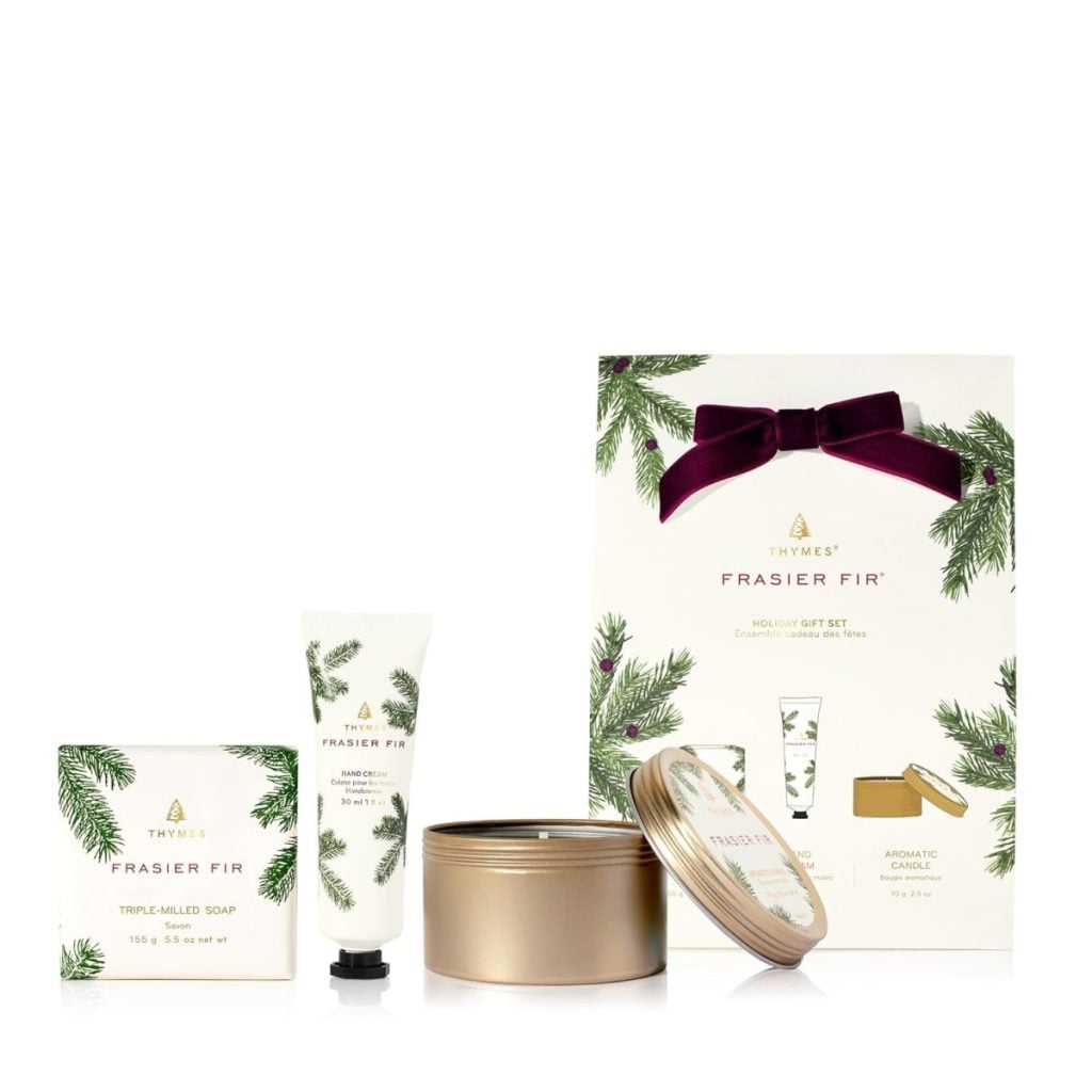 Gift set includes a bar soap, hand cream, and candle with Frasier Fir scent, displayed in front of decorated packaging with pine branch and ribbon design.