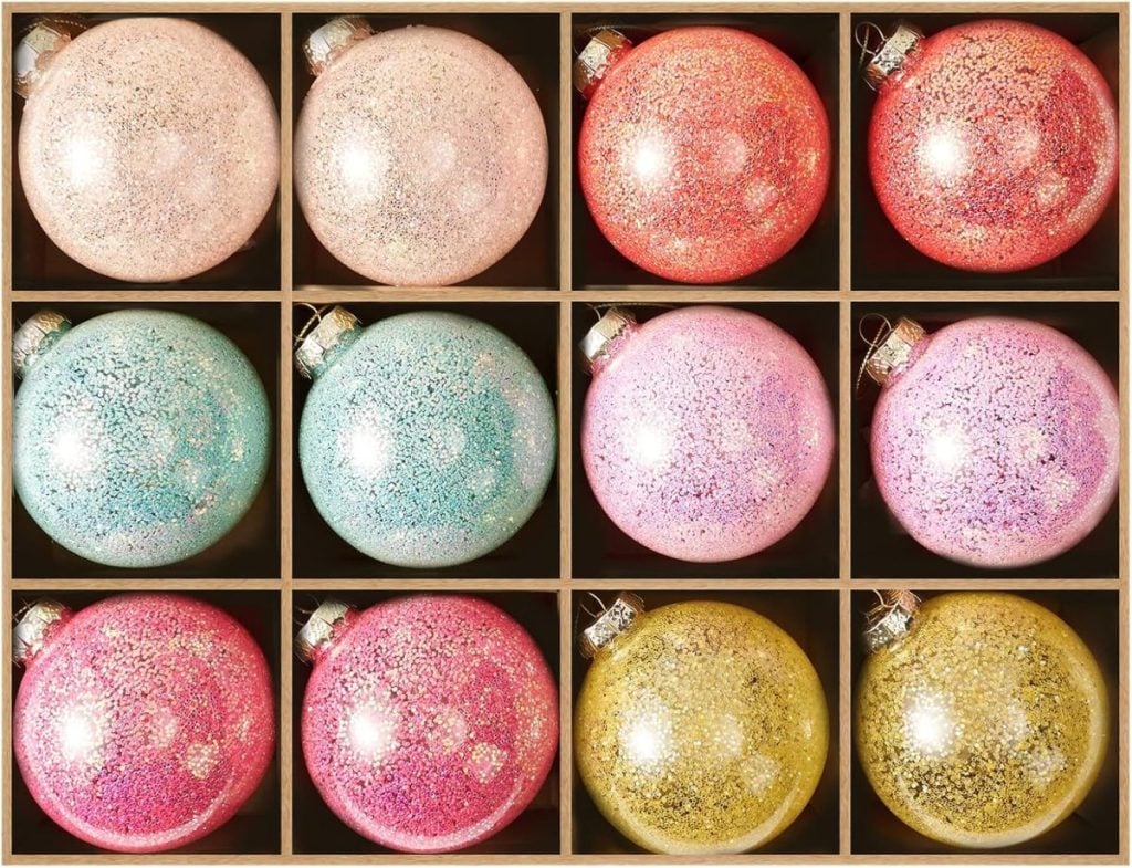 Twelve glittery Christmas ornaments in pastel shades are arranged in a wooden grid, each ornament placed in its own compartment.