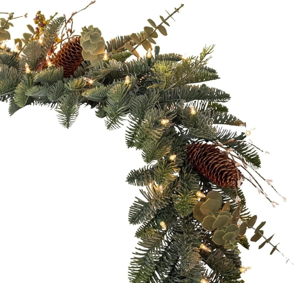 A close-up of a Christmas wreath decorated with pinecones, eucalyptus leaves, and string lights against a white background.