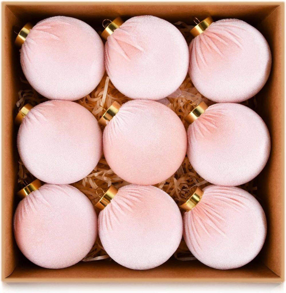 A box of nine matte pink Christmas ornaments with gold caps, arranged in a three-by-three grid on a bed of shredded paper.
