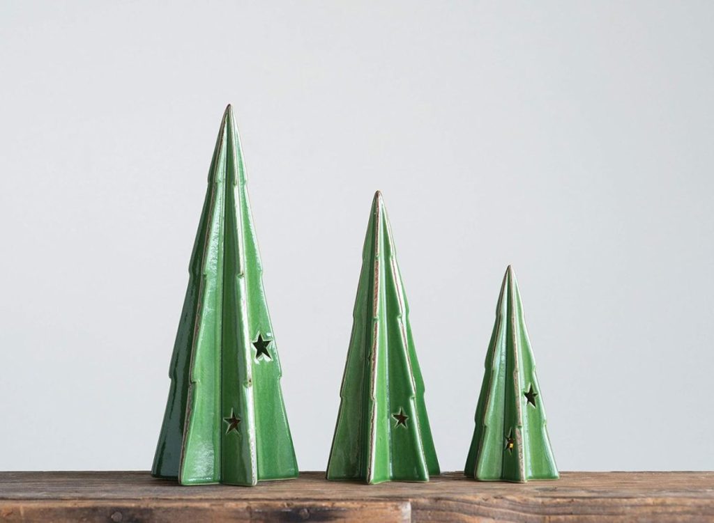 Three green ceramic Christmas tree decorations of varying heights with star-shaped cutouts are displayed on a wooden surface against a plain background.