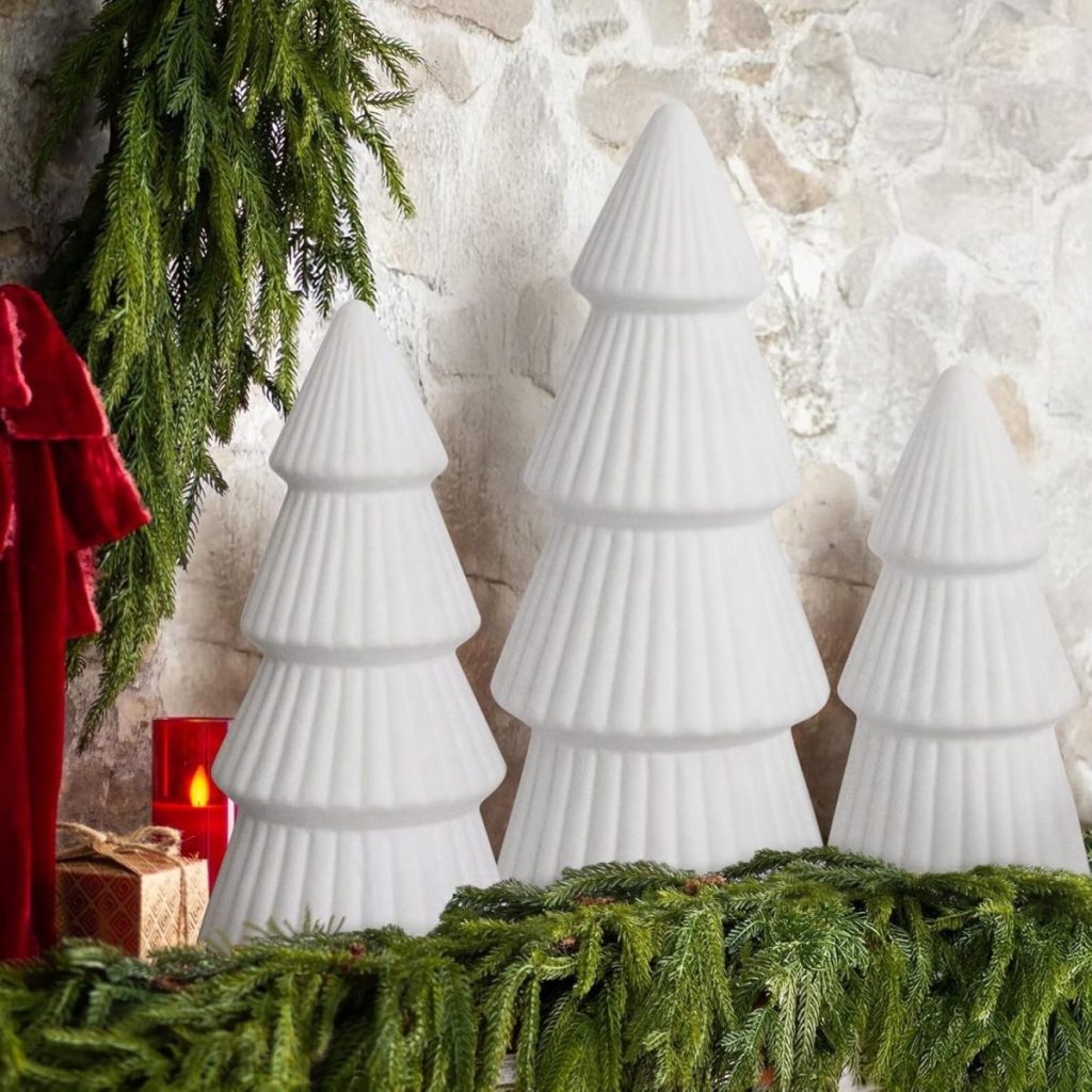 Three white, stylized ceramic Christmas trees are arranged in front of a stone wall, with green garland, a red candle, and evergreen branches surrounding them.