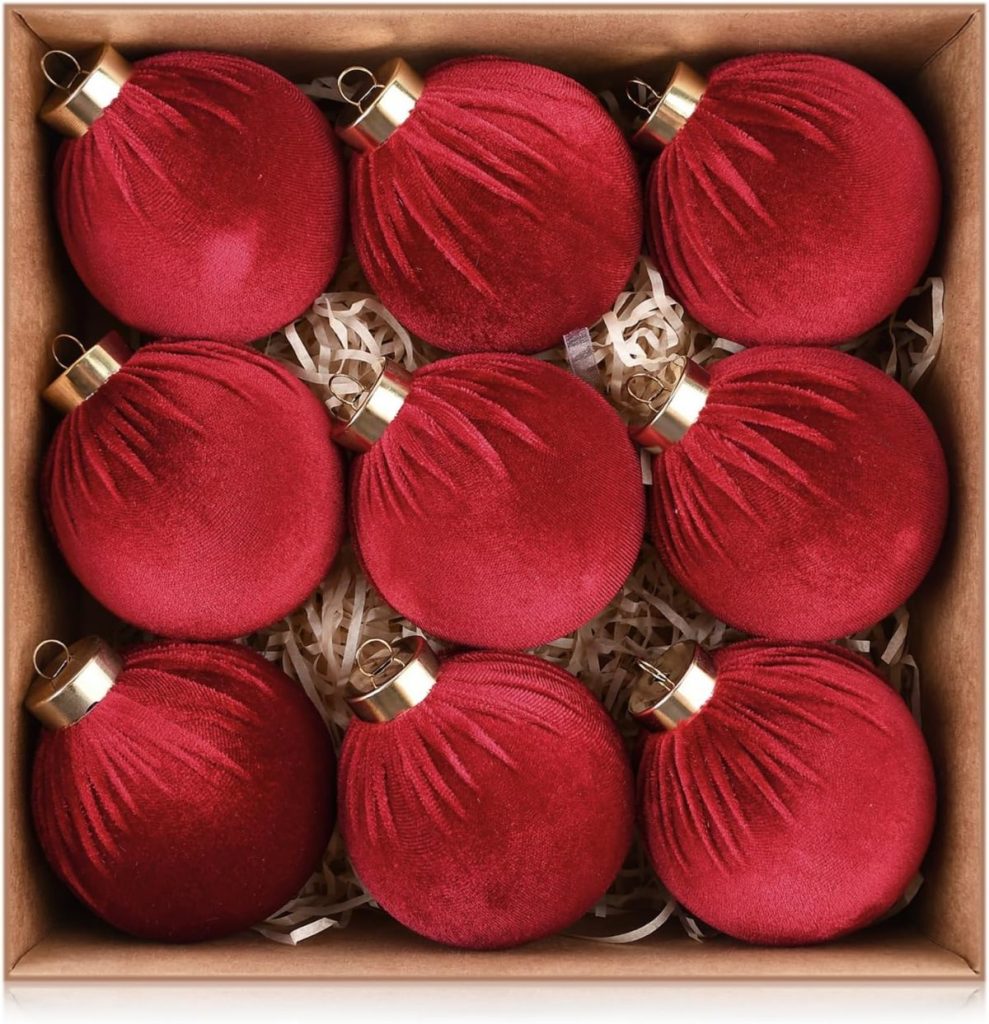 A box containing nine red velvet Christmas ornaments arranged in three rows on a bed of paper shreds.