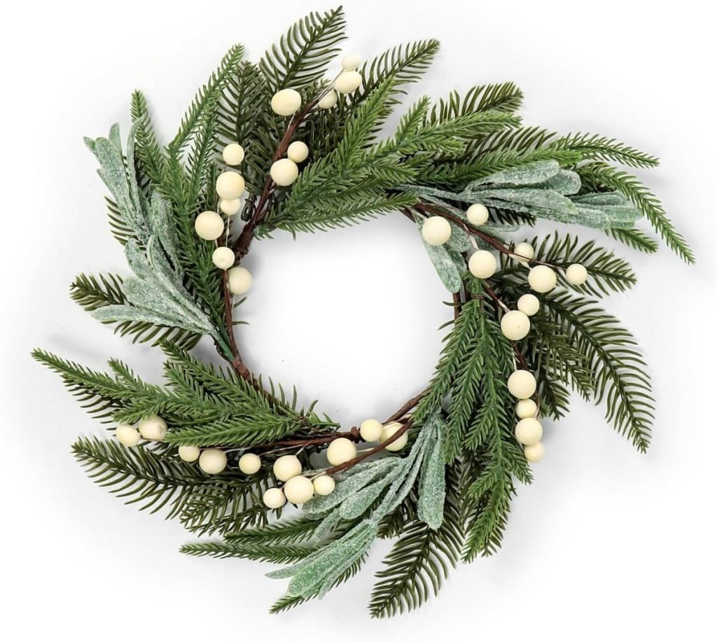 A circular wreath made of green pine branches, frosted leaves, and white berries arranged on a white background.