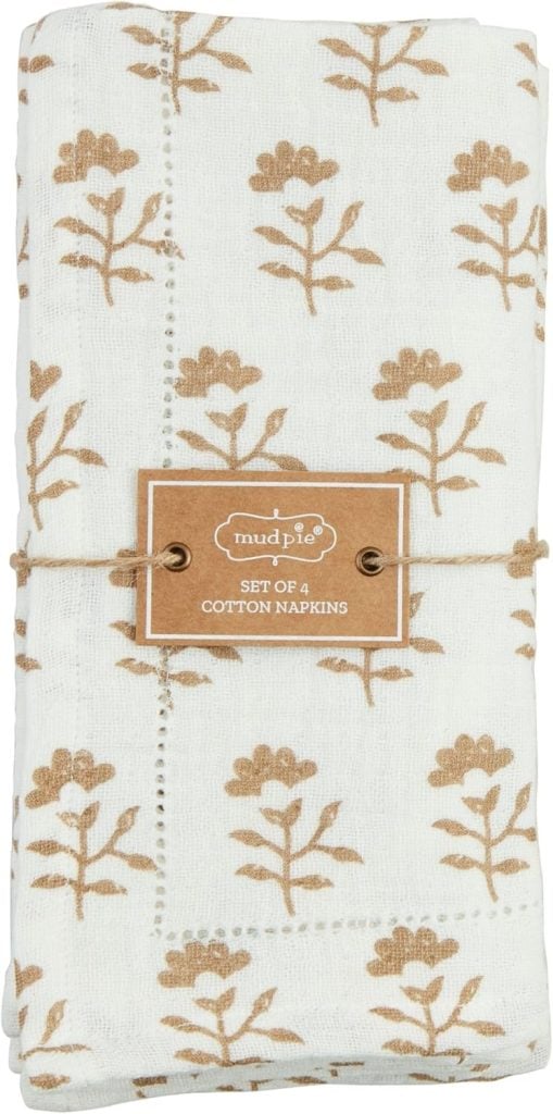 Folded white cotton napkins with a brown floral pattern, wrapped in twine with a label that reads "mud pie, set of 4 cotton napkins.