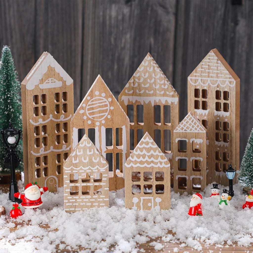 Wooden cutouts shaped like houses with white painted details are arranged on fake snow, surrounded by miniature Christmas figures, trees, and lampposts.