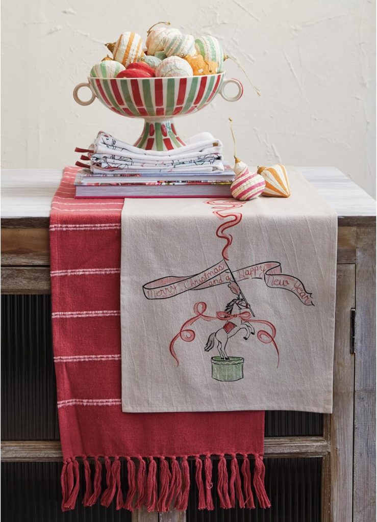 A table with stacked dish towels, a bowl of fabric pumpkins, and two decorative cloths draped over the edge, one red and one with a "Merry Christmas Happy New Year" unicorn design.