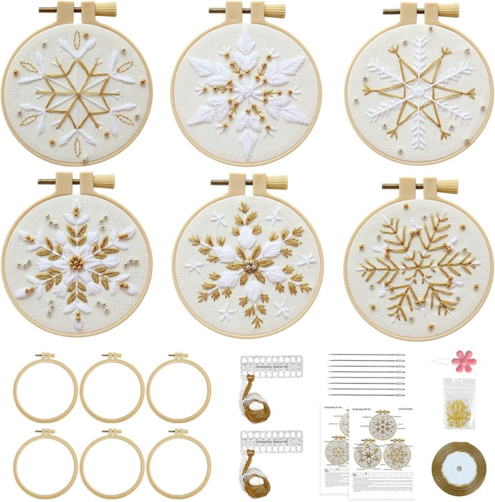 A set of six embroidery hoops with gold and white snowflake designs, embroidery threads, needles, instructions, and additional accessories arranged below the hoops.