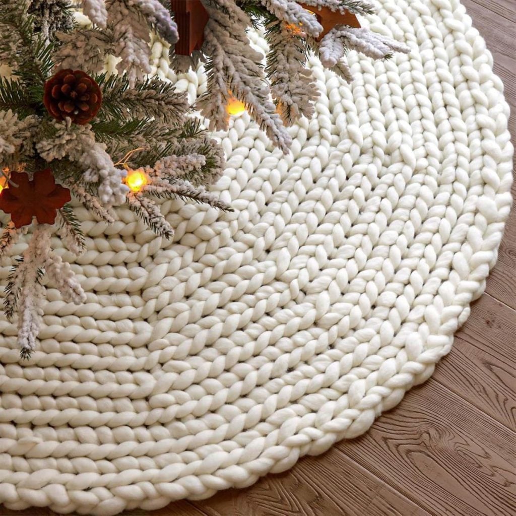 A round, chunky-knit cream tree skirt is placed under a snow-dusted Christmas tree with pinecones and lights, on a wooden floor.