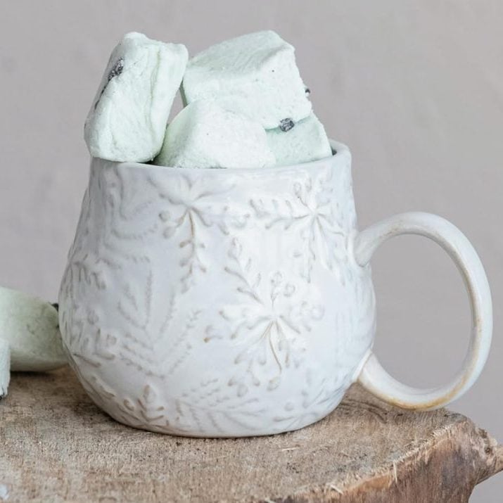 A white ceramic mug with an embossed leaf pattern is filled with pale green marshmallows, set on a wooden surface beside scattered treats—perfectly capturing cozy vibes and inspiring window art creativity.