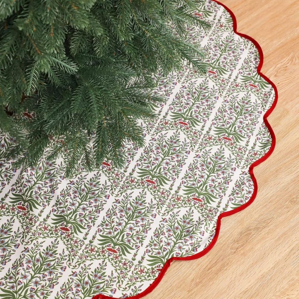 A Christmas tree sits on a patterned tree skirt with red trim, featuring green foliage and small red birds, on a wooden floor.